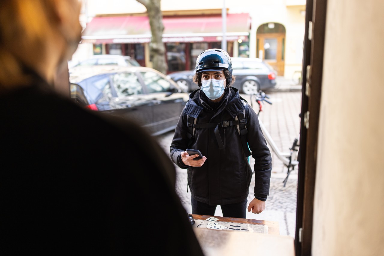 Food courier standing at resturant front for picking up take-out food orders during the coronavirus pandemic. Delivery person wearing face mask standing at restaurant takeaway counter.