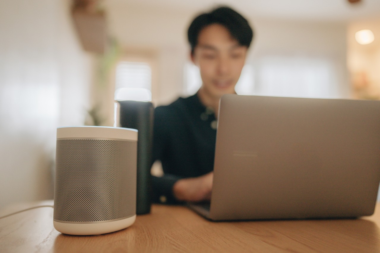 Young Asian Man Sitting at His Desk Works on Laptop, Beside Him Smart Speaker with AI Assistance Answers Questions, Plays Music, Podcast.